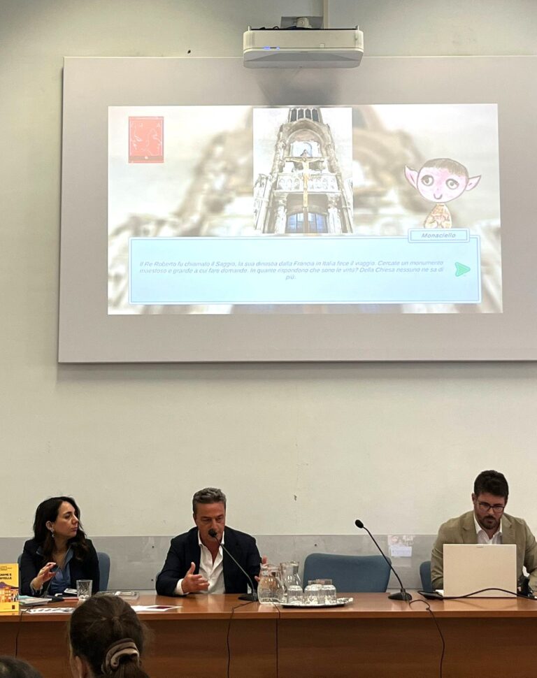 Il CEO di L’ABCD Edutainment Nicola D’Ambrosio parla al microfono durante un convegno, seduto tra due relatori. Sullo schermo alle sue spalle è proiettata un’immagine con un avatar chiamato “Monaciello”, parte del progetto AI presentato durante l’evento.